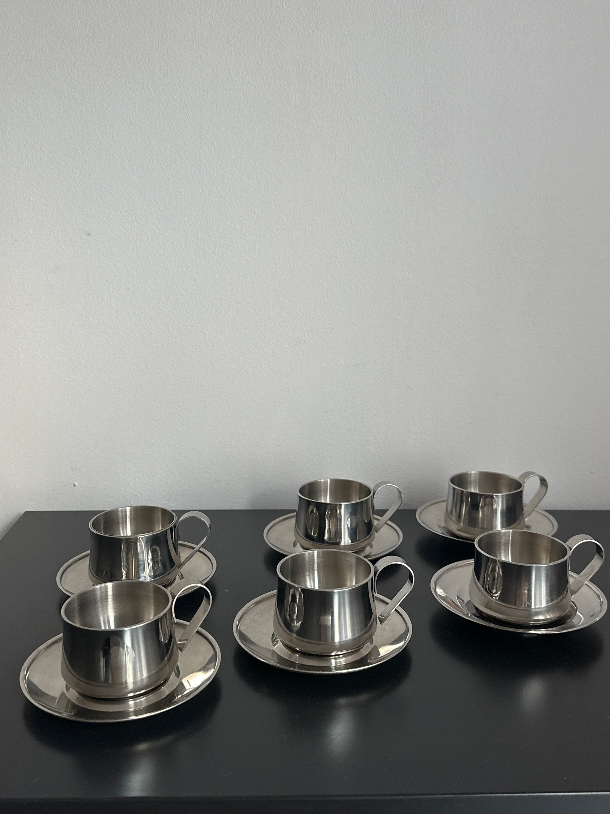 Three stainless steel cup holders with matching saucers mounted on a black wall-mounted panel against a light gray wall.