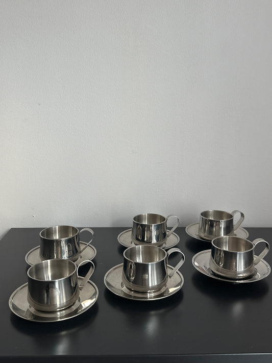 Three stainless steel cup holders with matching saucers mounted on a black wall-mounted panel against a light gray wall.