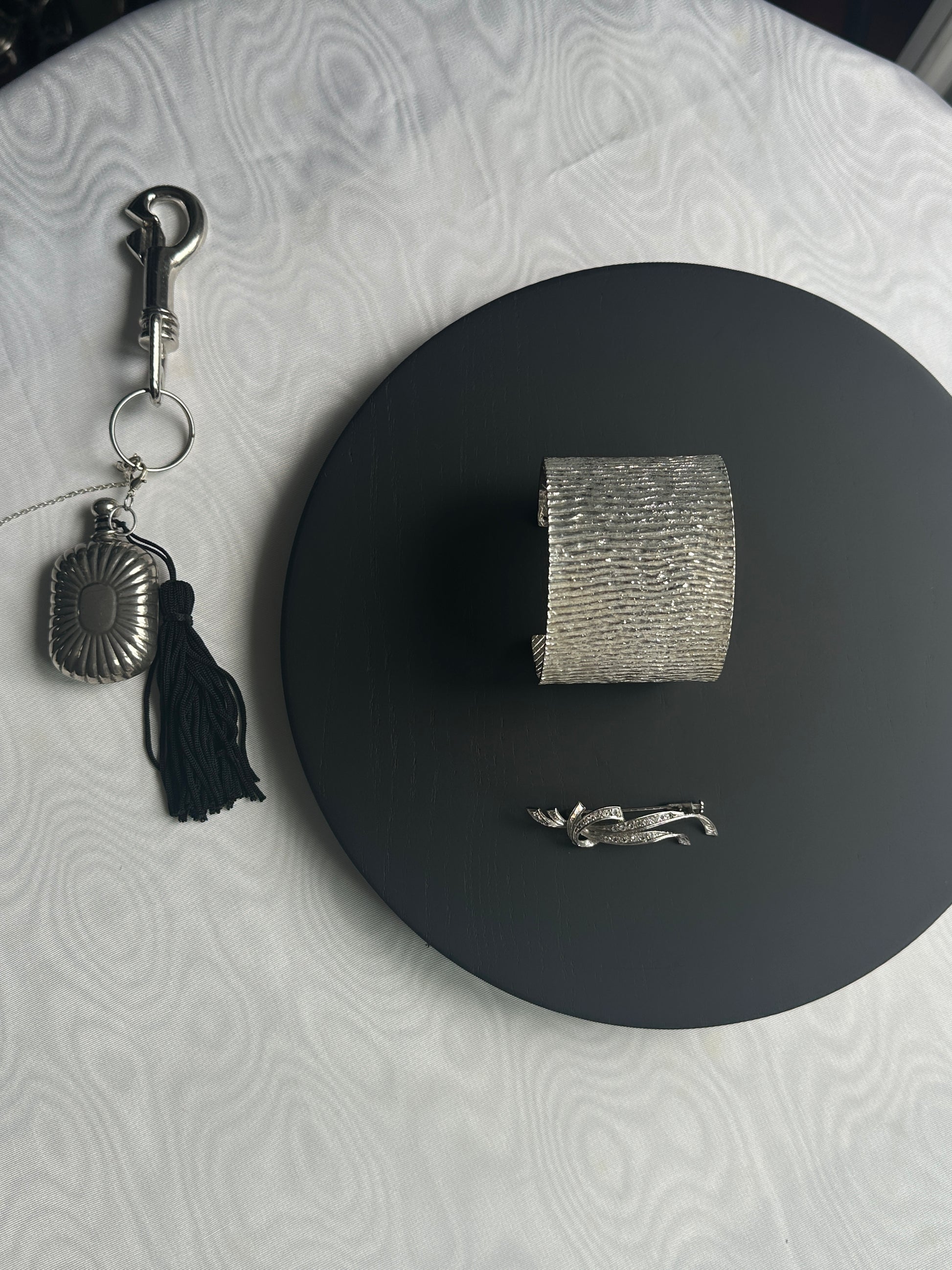 Silver bracelet and vintage perfume bottle  on a black circular mat with a textured moiree fabric background