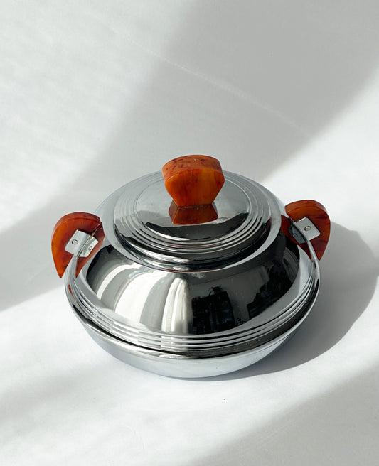 A polished stainless steel lidded box or sugar bowl with two reddish-brown side handles and a matching lid handle, photographed on a white surface with strong sunlight and shadows.